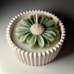 May include: A decorative candle shaped like a daisy flower, in shades of green and white, sits in a light pink, ridged container. The candle has a central wick and is designed to resemble a flower. The container has a scalloped edge.