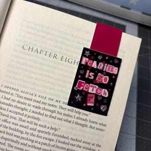 May include: A magnetic bookmark with a dark background featuring the pink text Reading is SO Fetch, surrounded by small pink hearts, stars, and a small book illustration. The bookmark is clipped onto the page of an open book showing Chapter Eight.