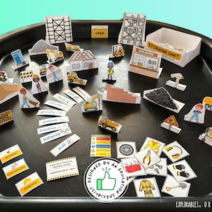 May include: A collection of paper construction-themed toys, including buildings, vehicles, workers, and signs, arranged on a black tray. The set includes a storage unit, traffic lights, and labels with words like "force" and "gravity". The design is labeled "EXPLORABLES. ORIGINAL."