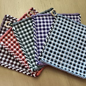 May include: Five woven pot holders in different colors and patterns. The pot holders are made of a woven fabric with a checkered pattern. The pot holders have a wooden ring attached to the top.