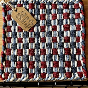 May include: A woven pot holder with a red, white, and blue checkered pattern. The pot holder is handwoven in the USA and made of 100% cotton. The tag attached to the pot holder reads "Kristin Kays - LOOPS ON A LOOM - handwoven in the USA - 100% cotton - machine wash - dry flat."