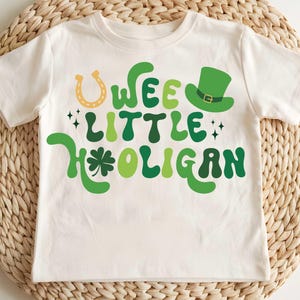 May include: White t-shirt with green and gold text that reads "Wee Little Hooligan". The text is in a retro font style. There is a green shamrock and a gold horseshoe above the text. A green leprechaun hat with a gold buckle is above the text.