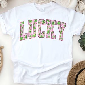May include: White t-shirt with the word "LUCKY" printed in pink letters. The letters are filled with a pattern of green shamrocks.