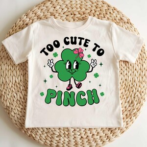 May include: White t-shirt with a green shamrock graphic and the text "Too Cute To Pinch".