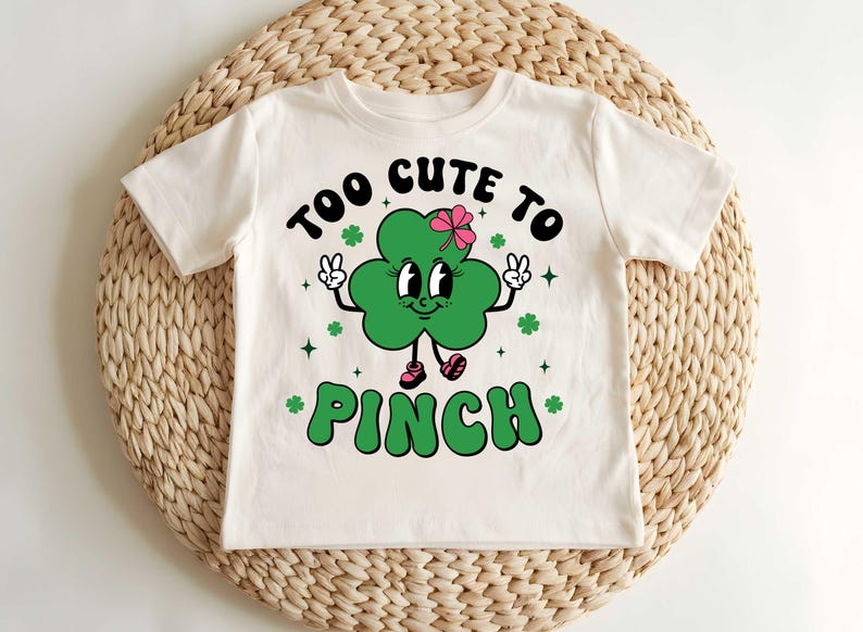 May include: White t-shirt with a green shamrock graphic and the text "Too Cute To Pinch".