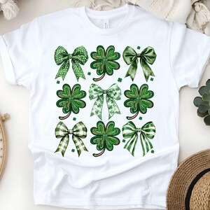 May include: White t-shirt featuring a green shamrock and bow design. The shamrocks and bows are green with white polka dots.