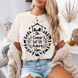 Pu&ograve; includere: Una maglietta bianca con un design grafico nero che rappresenta una scena della nativit&agrave; e il testo "Oh Come Let Us Adore Him".