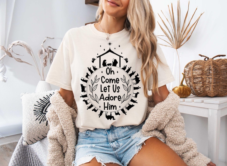 Pu&ograve; includere: Una maglietta bianca con un design grafico nero che rappresenta una scena della nativit&agrave; e il testo "Oh Come Let Us Adore Him".