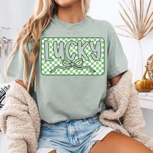 May include: A light green t-shirt with a green and white checkered box that says "LUCKY" in white letters with a green shamrock bow.