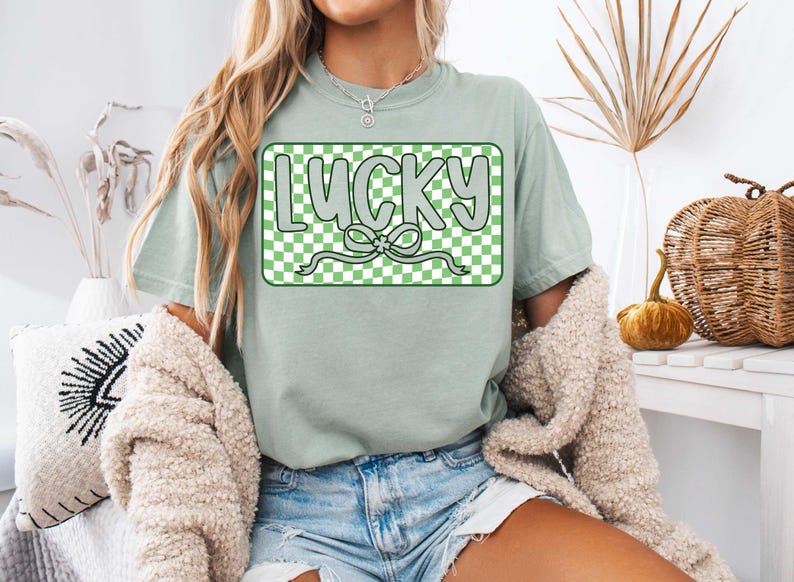 May include: A light green t-shirt with a green and white checkered box that says "LUCKY" in white letters with a green shamrock bow.