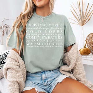 Pu&ograve; includere: Una maglietta verde chiaro con testo bianco che recita "Christmas Movies, Crackling Fires, Cold Noses, Frosty Mornings, Comfy Sweaters, Sparkling Snow, Warm Cookies, Gingerbread Candy Canes".