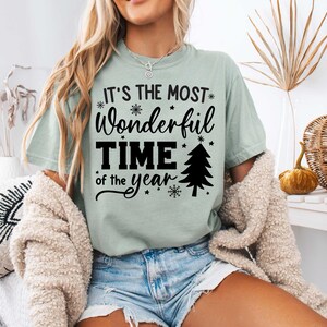 Pu&ograve; includere: Una maglietta verde chiaro con il testo "It's the most wonderful time of the year" in nero. Il testo &egrave; circondato da fiocchi di neve e stelle. Un piccolo albero di Natale &egrave; incluso anche nel design.