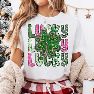 May include: A white t-shirt featuring a green four-leaf clover and the word "LUCKY" repeated three times in pink, green and white. The letters are outlined in black and have a polka dot pattern.