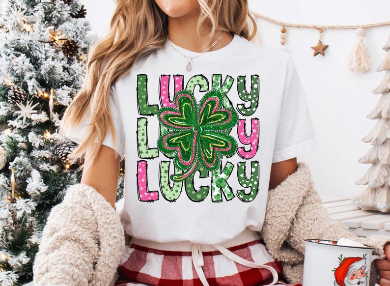 May include: A white t-shirt featuring a green four-leaf clover and the word "LUCKY" repeated three times in pink, green and white. The letters are outlined in black and have a polka dot pattern.
