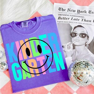 K&ouml;nnte beinhalten: Ein lila T-Shirt mit dem Wort "KINDERGARTEN" in bunten Buchstaben und einem Smiley-Gesicht. Das Shirt liegt auf einer wei&szlig;en Oberfl&auml;che, im Hintergrund eine Zeitung und eine Discokugel.