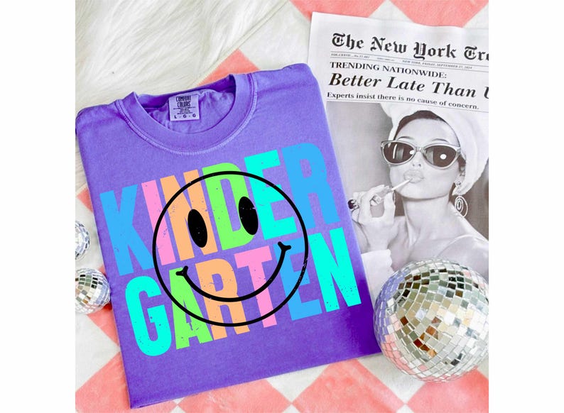 K&ouml;nnte beinhalten: Ein lila T-Shirt mit dem Wort "KINDERGARTEN" in bunten Buchstaben und einem Smiley-Gesicht. Das Shirt liegt auf einer wei&szlig;en Oberfl&auml;che, im Hintergrund eine Zeitung und eine Discokugel.