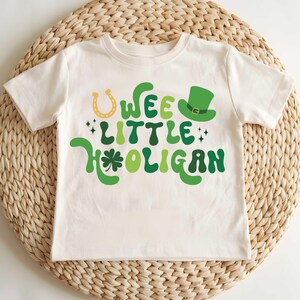 May include: A white t-shirt with a green, yellow, and black graphic that says "Wee Little Hooligan" with a shamrock and horseshoe.