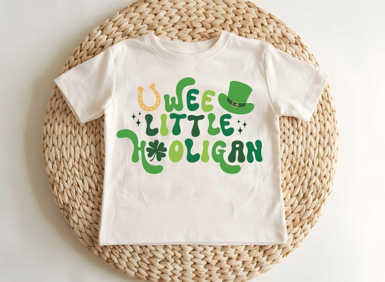 May include: A white t-shirt with a green, yellow, and black graphic that says "Wee Little Hooligan" with a shamrock and horseshoe.