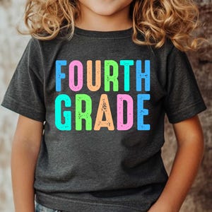 Op de afbeelding: Een donkergrijs t-shirt met de woorden "FOURTH GRADE" in grote, kleurrijke, verweerde letters. De tekst is in tinten blauw, roze, oranje en groen. Het shirt wordt door een kind gedragen.