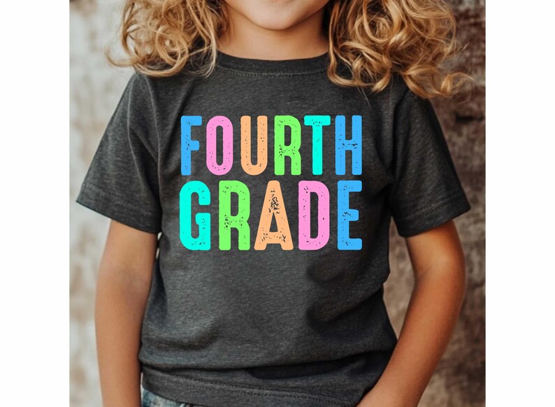 Op de afbeelding: Een donkergrijs t-shirt met de woorden "FOURTH GRADE" in grote, kleurrijke, verweerde letters. De tekst is in tinten blauw, roze, oranje en groen. Het shirt wordt door een kind gedragen.