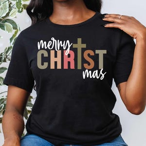 Pu&ograve; includere: Una maglietta nera con il testo "merry CHRISTmas" in lettere colorate. La parola "CHRIST" &egrave; in un carattere pi&ugrave; grande rispetto alle altre parole. Una croce &egrave; centrata sopra la parola "CHRIST".