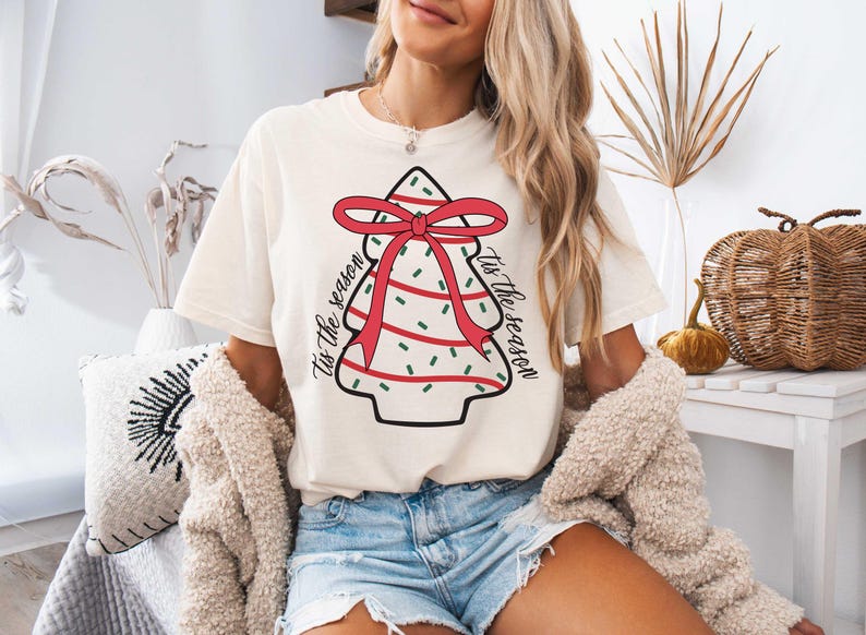 Pu&ograve; includere: Una maglietta bianca con un design di albero di Natale rosso e verde. L'albero ha un fiocco rosso e il testo "Tis the season" &egrave; scritto su entrambi i lati dell'albero.