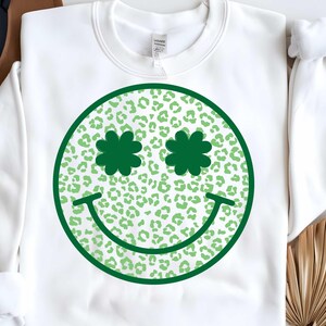 May include: White sweatshirt with a green smiley face design. The smiley face has shamrock-shaped eyes and a green leopard print pattern.