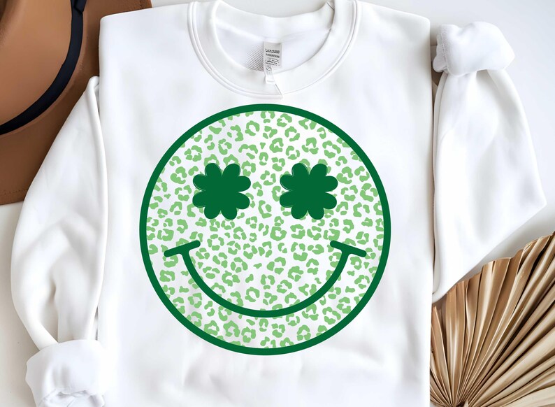 May include: White sweatshirt with a green smiley face design. The smiley face has shamrock-shaped eyes and a green leopard print pattern.