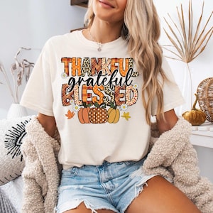 K&ouml;nnte beinhalten: Ein wei&szlig;es T-Shirt mit einem grafischen Design, das "thankful grateful blessed" in einem Patchwork-Stil mit Herbstfarben und K&uuml;rbissen sagt.