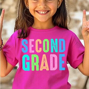 Op de afbeelding: Een magenta T-shirt met de woorden "SECOND GRADE" in kleurrijke, versleten letters. Het shirt wordt gedragen door een jong persoon die lacht en twee vingers omhoog houdt.