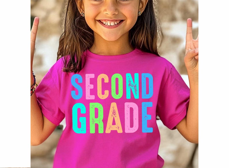 Op de afbeelding: Een magenta T-shirt met de woorden "SECOND GRADE" in kleurrijke, versleten letters. Het shirt wordt gedragen door een jong persoon die lacht en twee vingers omhoog houdt.