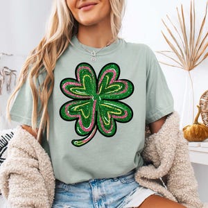 May include: A light green t-shirt featuring a green, pink and yellow glitter four-leaf clover design.