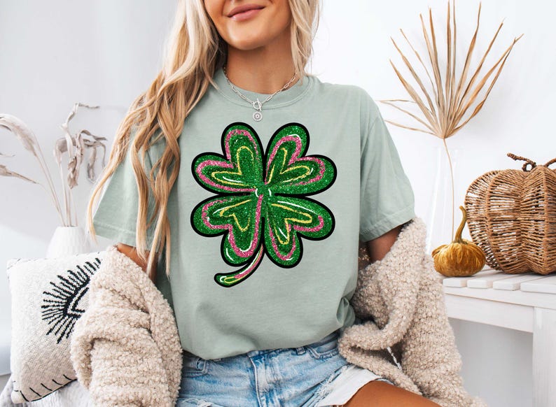 May include: A light green t-shirt featuring a green, pink and yellow glitter four-leaf clover design.