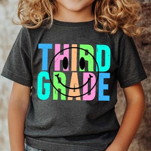 K&ouml;nnte beinhalten: Dunkelgraues T-Shirt mit den Worten "THIRD GRADE" in Blau, Rosa, Gr&uuml;n und Orange, mit einem schwarzen Smiley. Das T-Shirt wird von einem Kind getragen.