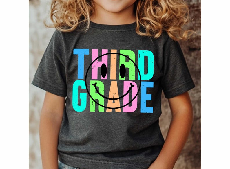 K&ouml;nnte beinhalten: Dunkelgraues T-Shirt mit den Worten "THIRD GRADE" in Blau, Rosa, Gr&uuml;n und Orange, mit einem schwarzen Smiley. Das T-Shirt wird von einem Kind getragen.