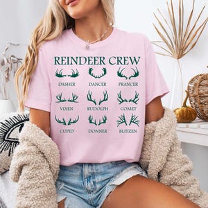 Pu&ograve; includere: Maglietta rosa con il testo "Reindeer Crew" e una grafica di corna di renna con i nomi di ogni renna: Dasher, Dancer, Prancer, Vixen, Rudolph, Comet, Cupid, Donner, Blitzen.