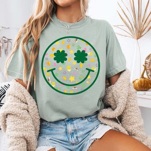 May include: A light green t-shirt featuring a green smiley face design. The smiley face has shamrock eyes and is filled with a pattern of shamrocks, stars, horseshoes, and rainbows.