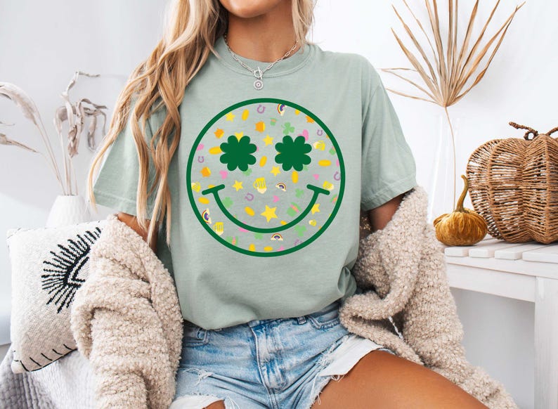 May include: A light green t-shirt featuring a green smiley face design. The smiley face has shamrock eyes and is filled with a pattern of shamrocks, stars, horseshoes, and rainbows.