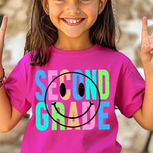 K&ouml;nnte beinhalten: Ein magentafarbenes T-Shirt mit den Worten "SECOND GRADE" in Blau, Rosa, Gr&uuml;n und Orange, mit einem schwarzen Smiley-Gesicht. Das T-Shirt wird von einer jungen Person getragen, die l&auml;chelt und das Friedenszeichen macht.