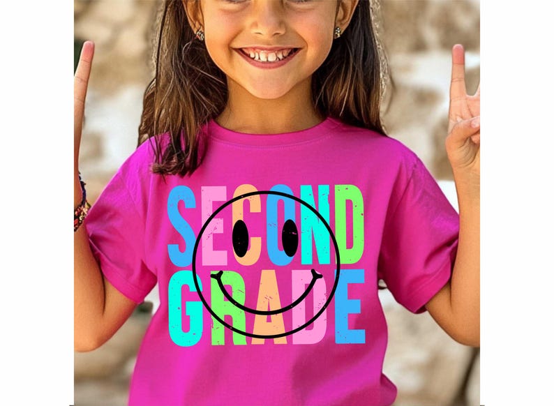 K&ouml;nnte beinhalten: Ein magentafarbenes T-Shirt mit den Worten "SECOND GRADE" in Blau, Rosa, Gr&uuml;n und Orange, mit einem schwarzen Smiley-Gesicht. Das T-Shirt wird von einer jungen Person getragen, die l&auml;chelt und das Friedenszeichen macht.