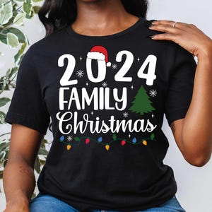 Pu&ograve; includere: Maglietta nera con testo bianco che recita "2024 Family Christmas" con un cappello di Babbo Natale rosso, un albero di Natale verde e luci di Natale colorate.