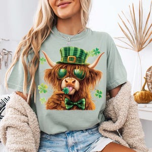 May include: A light green t-shirt featuring a cartoon image of a Highland cow wearing a green and white checked hat, sunglasses, and a green bow tie. The cow is holding a shamrock in its mouth.