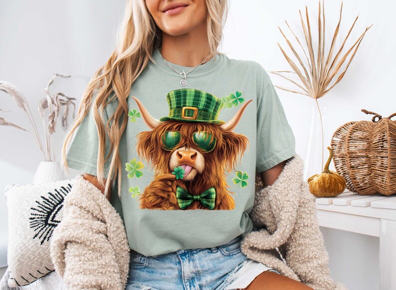 May include: A light green t-shirt featuring a cartoon image of a Highland cow wearing a green and white checked hat, sunglasses, and a green bow tie. The cow is holding a shamrock in its mouth.