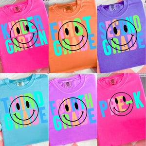 Puede incluir: Una colección de camisetas coloridas con diseños de niveles escolares. Cada camiseta presenta una gran carita sonriente con el texto del nivel escolar, como "First Grade" o "Second Grade", en colores brillantes y contrastantes. Las camisetas son de varios colores.