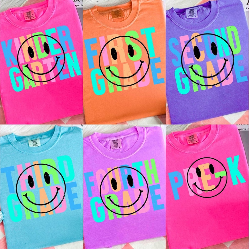 K&ouml;nnte beinhalten: Eine Sammlung farbenfroher T-Shirts mit Designs f&uuml;r verschiedene Klassenstufen. Jedes Shirt zeigt ein gro&szlig;es Smiley-Gesicht mit dem Text der Klassenstufe, wie z. B. "First Grade" oder "Second Grade", in leuchtenden, kontrastierenden Farben. Die Shirts sind in verschiedenen Farben.