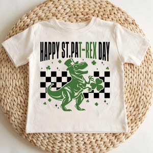 May include: White t-shirt with a green dinosaur wearing a top hat and holding a shamrock. The shirt says "Happy St. Pat-Rex Day".