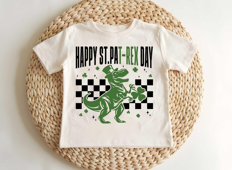 May include: White t-shirt with a green dinosaur wearing a top hat and holding a shamrock. The shirt says "Happy St. Pat-Rex Day".