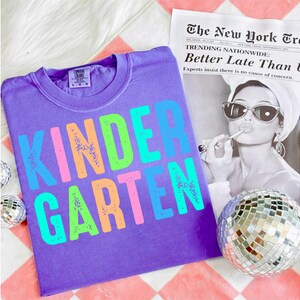 Op de afbeelding: Een paars T-shirt met het woord "KINDERGARTEN" in kleurrijke, verweerde letters. Het shirt wordt getoond met een zwart-wit afbeelding van een vrouw en een discobal. De krantenkop luidt "Better Late Than L".