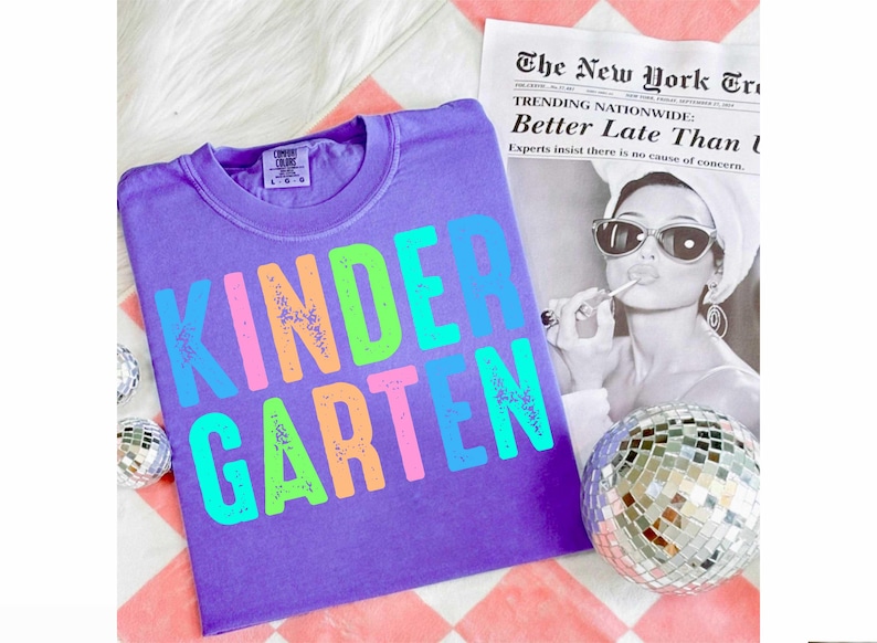 Op de afbeelding: Een paars T-shirt met het woord "KINDERGARTEN" in kleurrijke, verweerde letters. Het shirt wordt getoond met een zwart-wit afbeelding van een vrouw en een discobal. De krantenkop luidt "Better Late Than L".