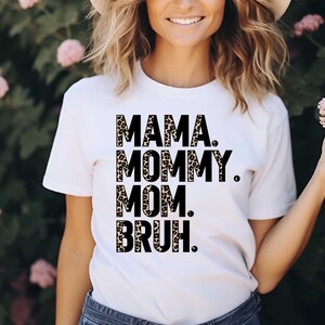 Puede incluir: Camiseta blanca con las palabras "MAMA. MOMMY. MOM. BRUH." impresas en negro y estampado de leopardo. La camiseta es de estilo casual, adecuada para el uso diario. El dise&ntilde;o es una versi&oacute;n divertida y moderna de la maternidad.